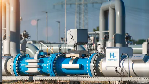 Um close-up de tubos industriais de aço em um ambiente externo, com um tubo azul central com flanges e uma placa branca rotulada "H2 HYDROGEN" em um tubo branco à direita. Um close-up de tubos industriais de aço em um ambiente externo, com um tubo azul central com flanges e uma placa branca rotulada "H2 HYDROGEN" em um tubo branco à direita.
